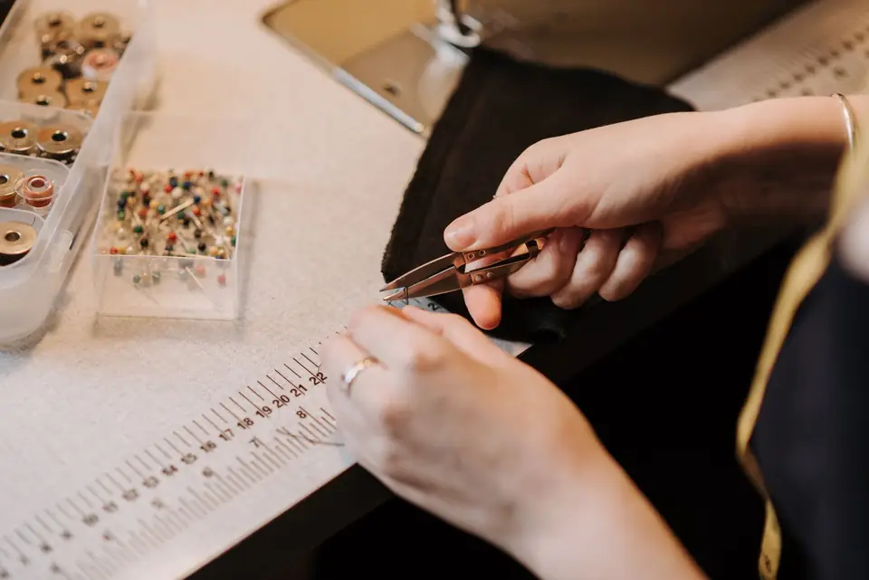 Dressmaker working on black fabric with sewing nippers, a ruler and a notions box nearby.