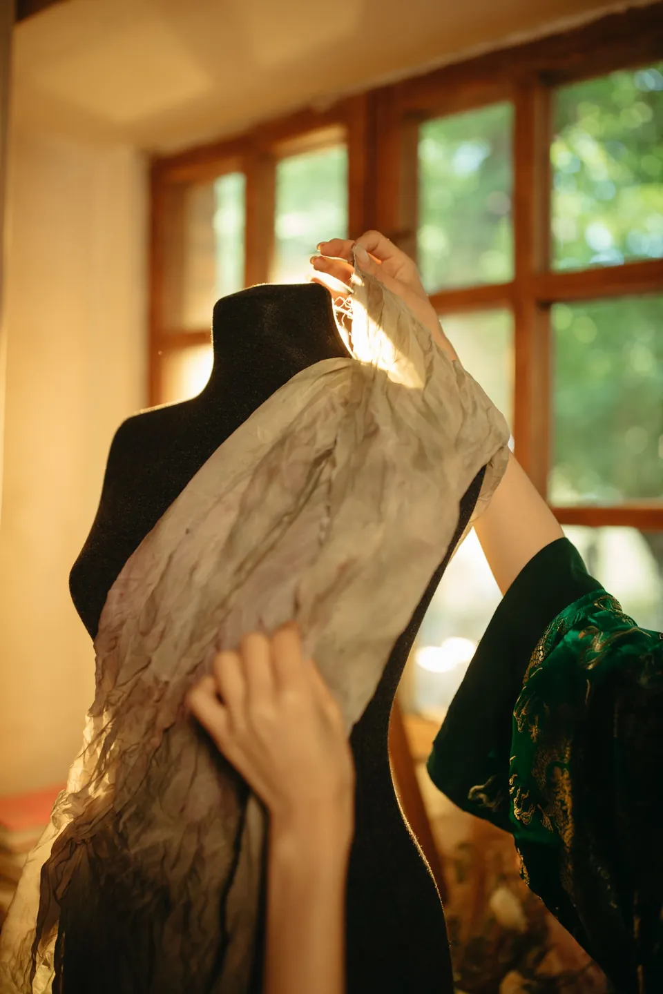 Hands draping a light fabric over a dress form in a sunlit atelier.