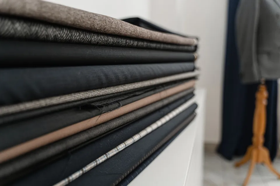 Stack of folded suiting fabrics, plain and patterned, in a tailoring workshop with a dress form behind.