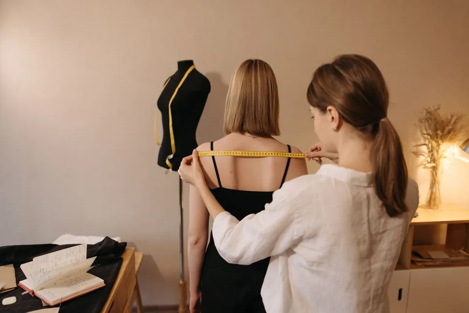 Dressmaker measuring a client’s shoulders with a tape measure in a workshop.