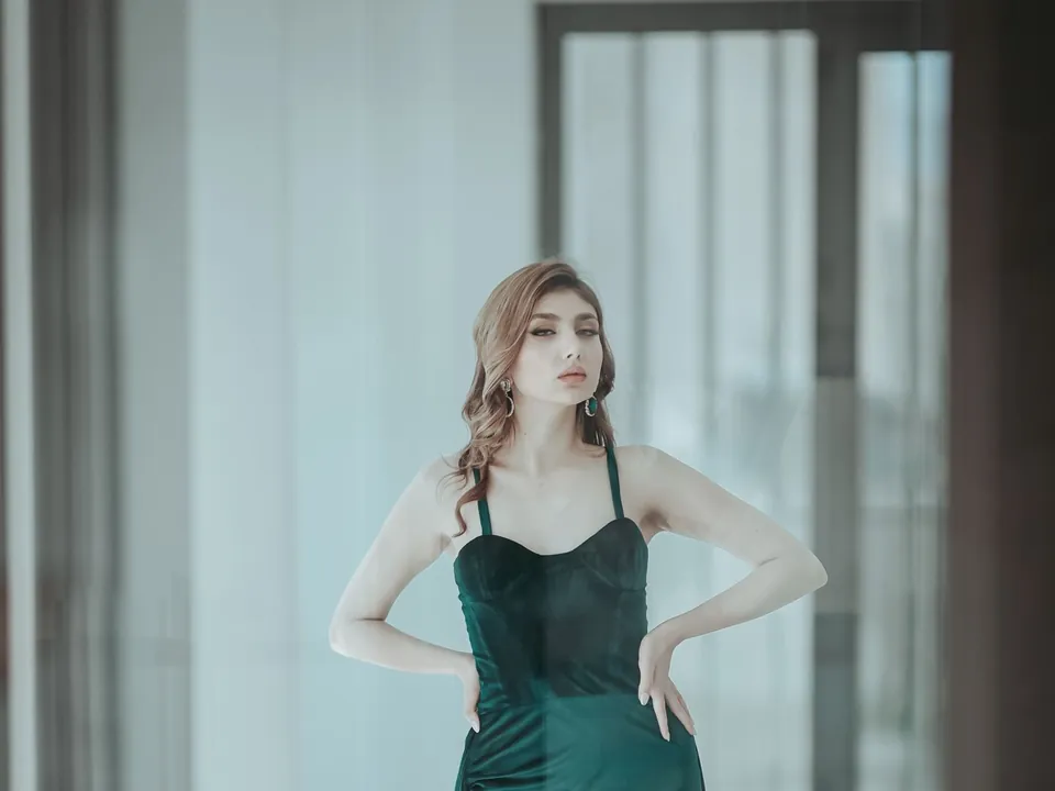 Woman in a long green satin dress with a slit, standing in a bright interior.