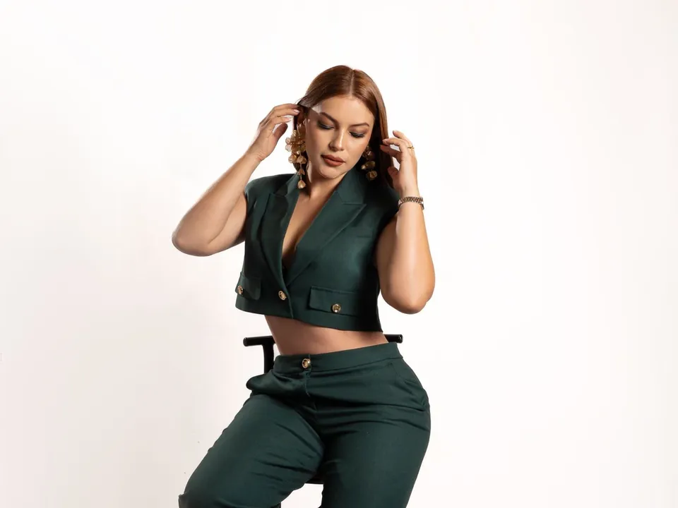 Woman in a green set, cropped jacket and trousers, sitting on a stool in a studio.