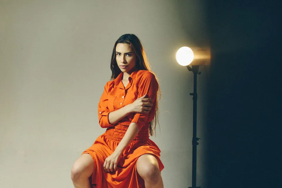 Woman in an orange shirt dress sitting on a stool, lit in a studio.