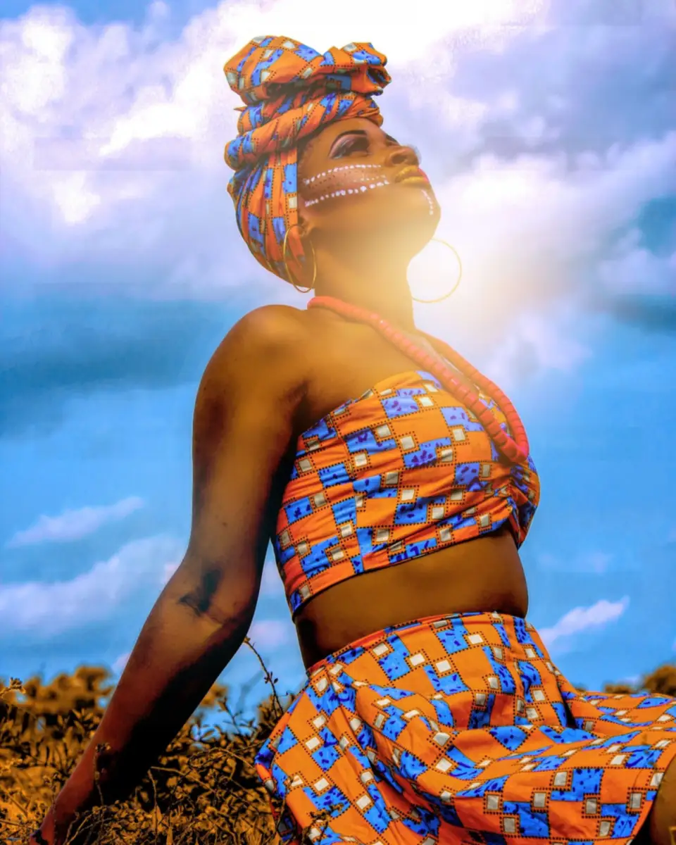 Woman in a head wrap wearing an orange and blue printed top and skirt, sitting in the sun.