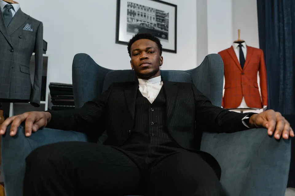 Man in a black three-piece suit seated in an armchair in a tailor’s workshop.