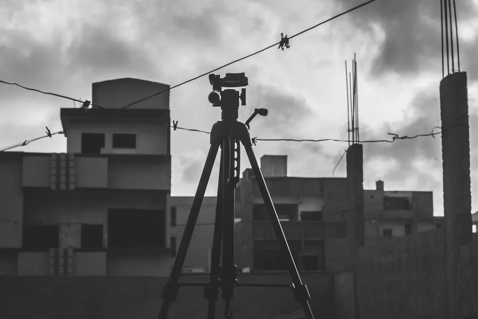 Camera tripod silhouetted on a rooftop facing unfinished buildings, black and white.