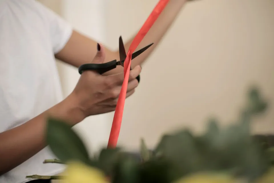 Hands cutting a red ribbon with scissors above foliage.