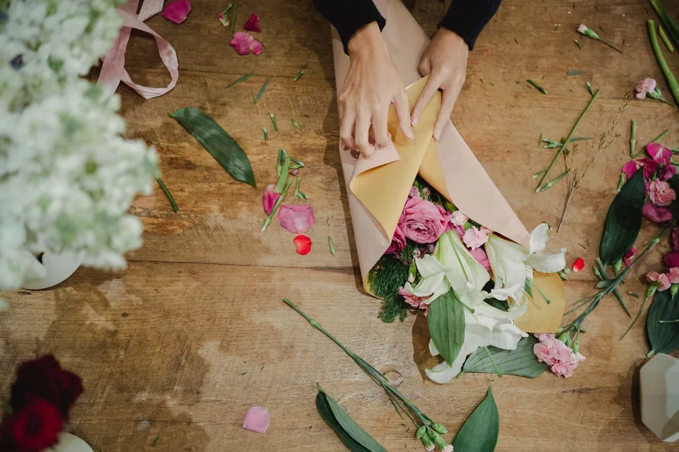 Hands wrapping a bouquet on a flower-strewn worktable, seen from above.