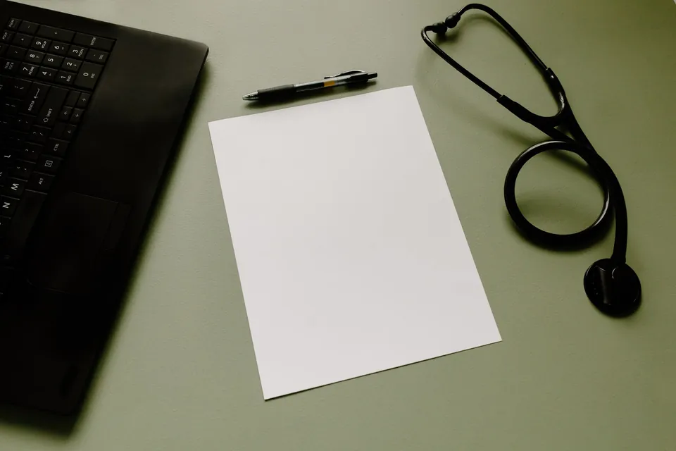 Stethoscope, pen, blank sheet, and laptop on a sage green desk.