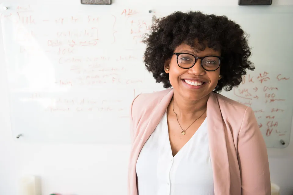 Smiling woman with glasses in a powder-pink jacket, in front of a whiteboard covered in notes.