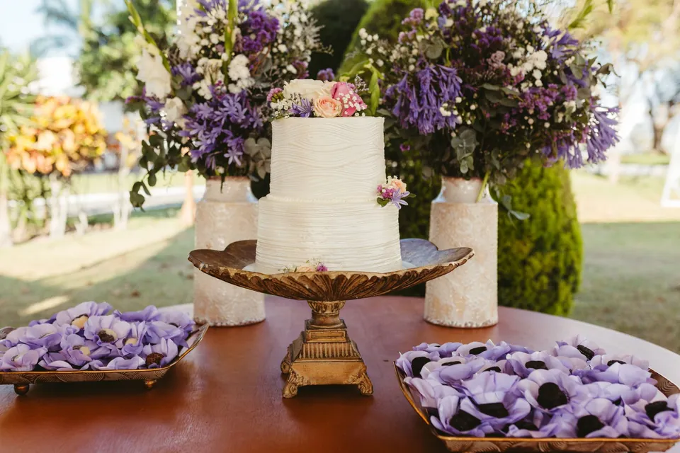 A white wedding cake surrounded by mauve flowers, set up outdoors.