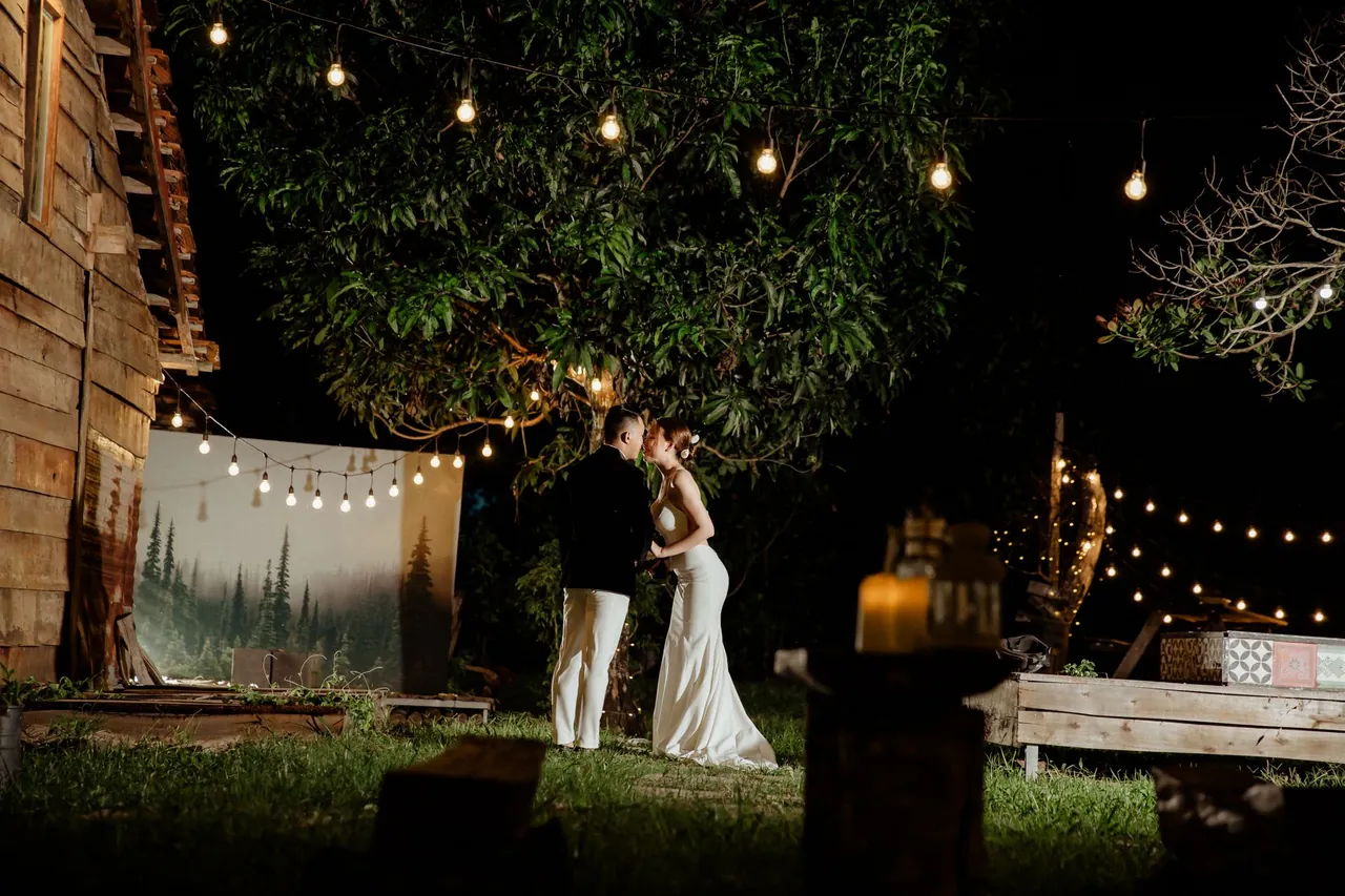 A wedding couple kisses under string lights in a garden at nightfall.