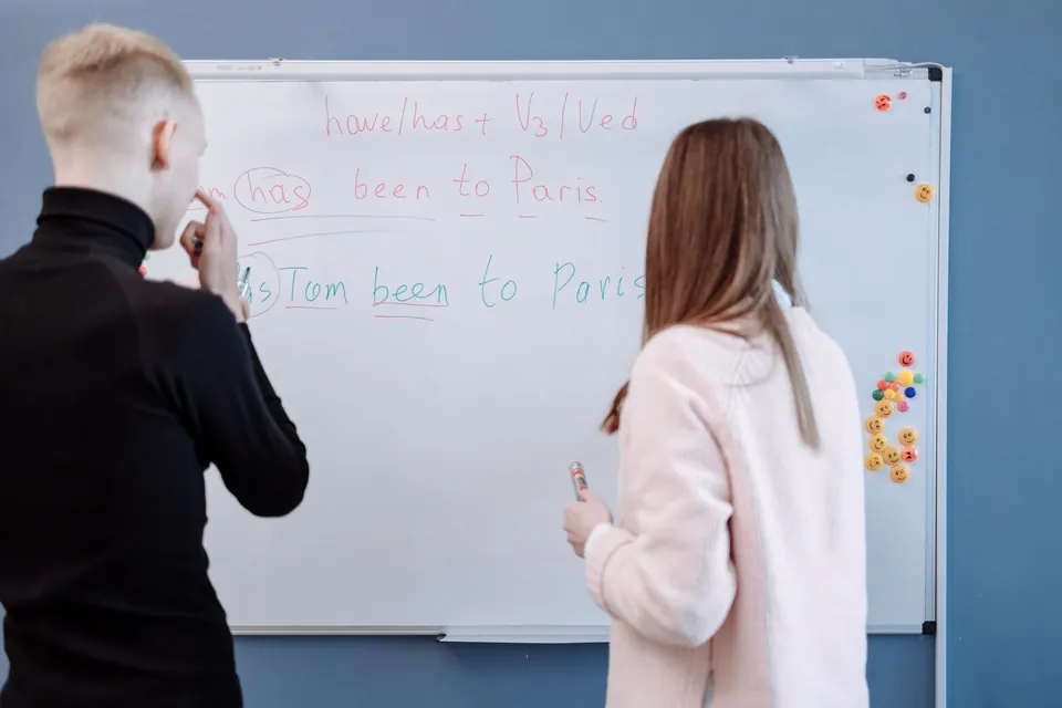 Two people, seen from behind, at a whiteboard covered with English sentences.