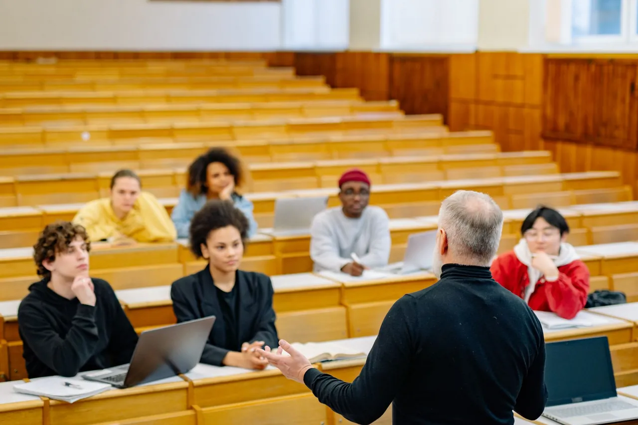 Teacher, seen from behind, addressing students seated in a lecture hall with wooden benches.