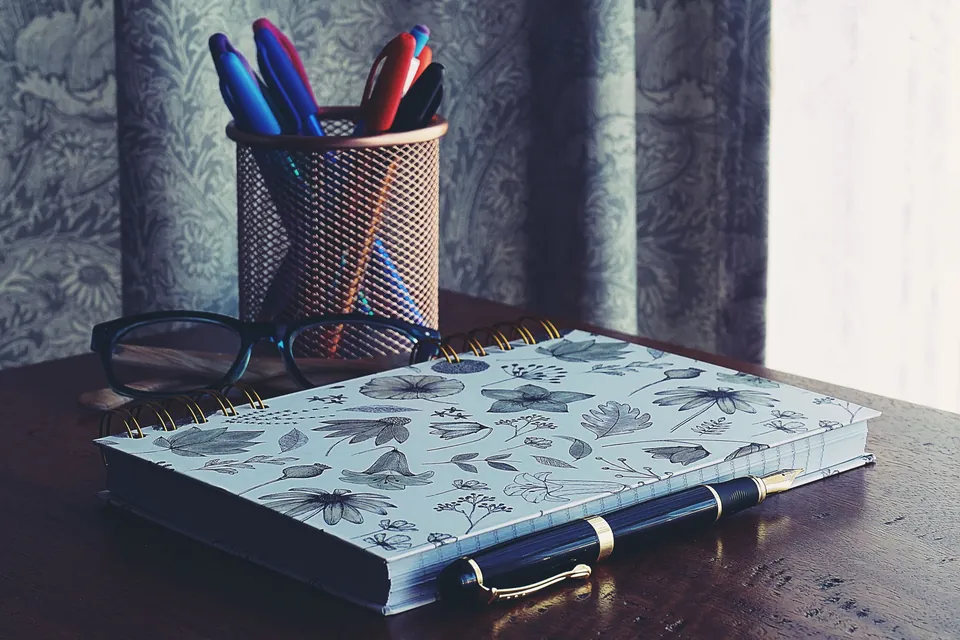 Spiral notebook, fountain pen, glasses and a pot of felt pens on a wooden desk.