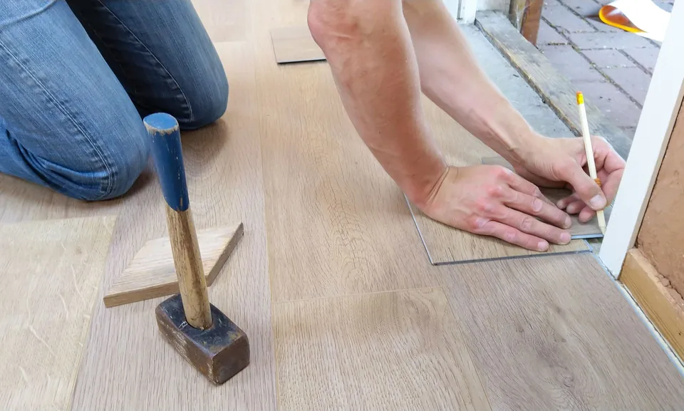 Craftsman kneeling to mark the cut of a floorboard, mallet beside him.