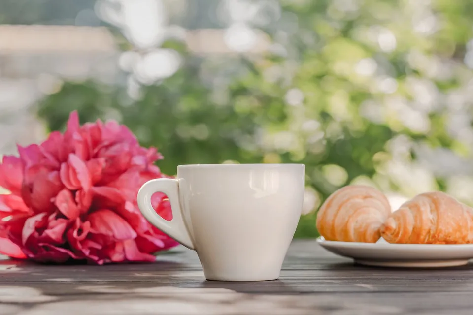 Cup of coffee, croissants and pink peonies on a wooden table.