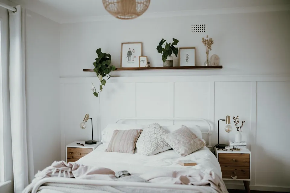 Bright bedroom: linen bed, wooden bedside tables and a wall shelf with plants and frames.