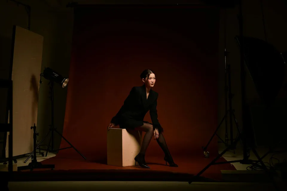 Studio portrait: woman in a black suit seated on a box against a lit brown backdrop.