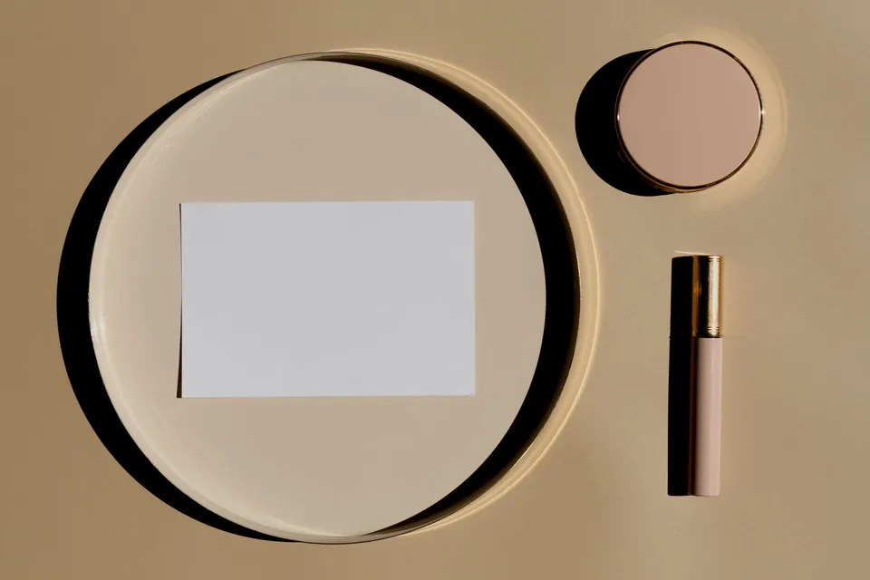 Top-down product set-up: blank card, compact powder and lipstick on a beige background.