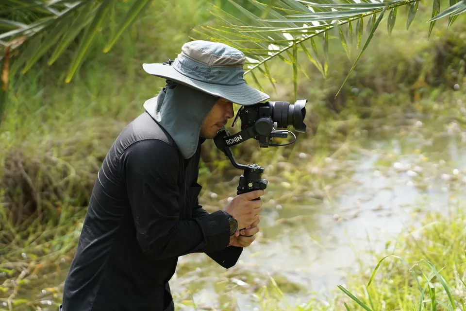 Videographer in a hat filming by the water with a stabiliser.