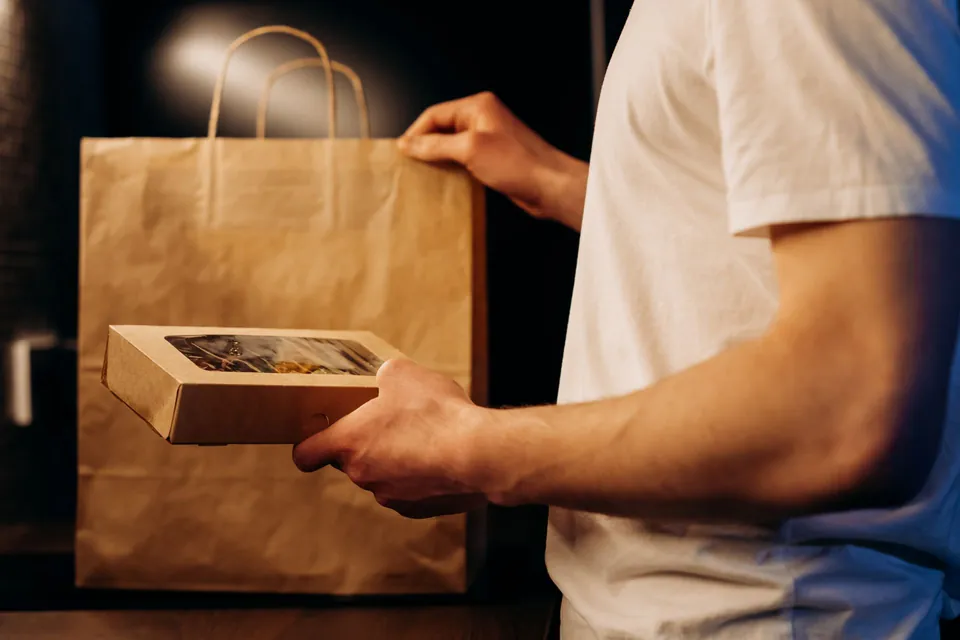 Hands slipping a cardboard meal box into a kraft bag for delivery.