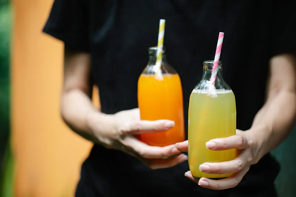 Hands holding two bottles of fresh juice, orange and yellow, with striped straws.