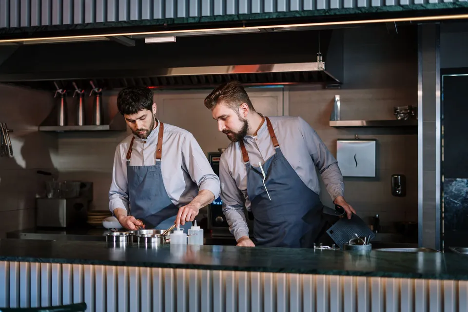 Two cooks in aprons preparing dishes in an open kitchen.