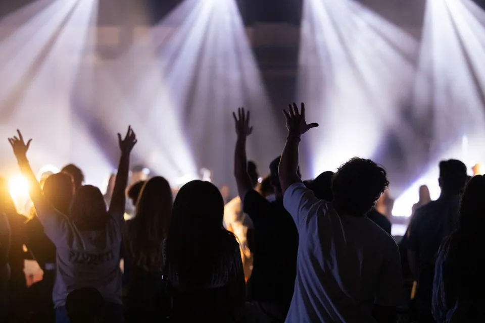 Audience with raised arms facing a stage flooded with white light.