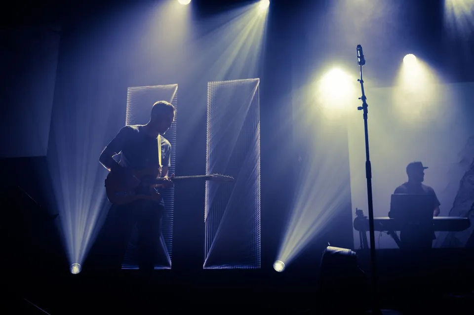 Guitarist in silhouette on a stage swept by blue light beams.