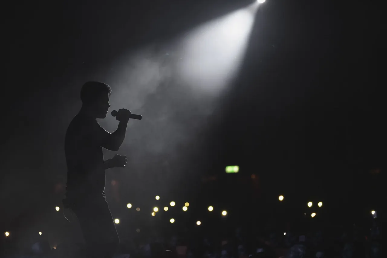 Silhouette of a performer singing into a microphone under a spotlight, in stage haze.